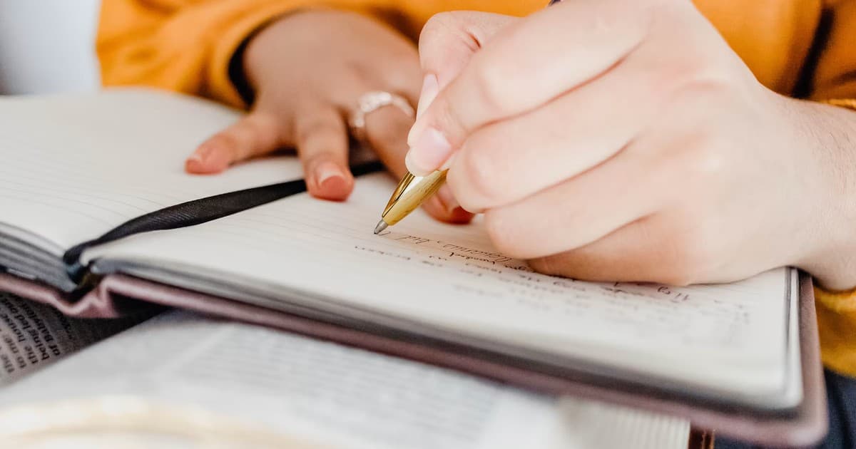 A person writes notes in an open notebook beside printed material.